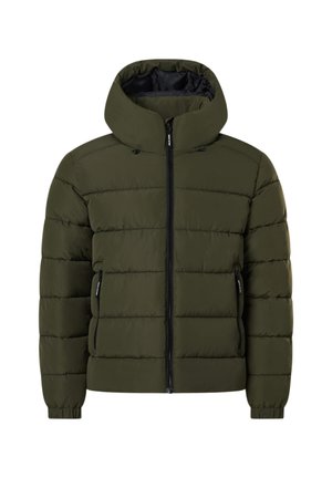 Jeune homme portant une doudoune vert olive, une chemise blanche, un pantalon noir et des bottes à lacets couleur sable, debout les mains dans les poches.