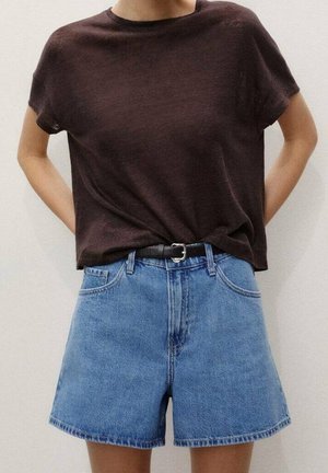 Person iført en mørkebrun kortærmet skjorte, stukket ned i højtaljede blå denimshorts med et sort bælte, stående mod en lys baggrund.