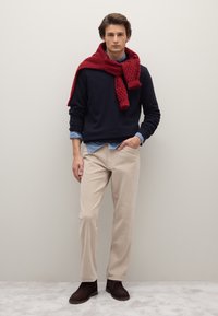 Suéter azul marino, camisa celeste, pantalones de pana beige y botas marrones. Bufanda roja texturizada caída sobre los hombros. Fondo neutro.