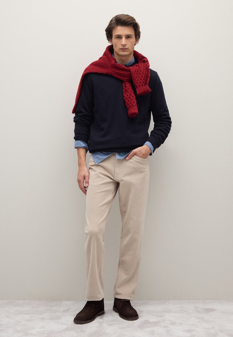 Suéter azul marino, camisa celeste, pantalones de pana beige y botas marrones. Bufanda roja texturizada caída sobre los hombros. Fondo neutro.