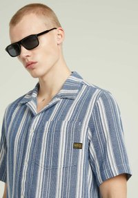 Korte mouwen, knoopsluiting shirt met blauwe en witte verticale strepen, een borstzak en een merklogo-label. Vergezeld van zwarte zonnebril.