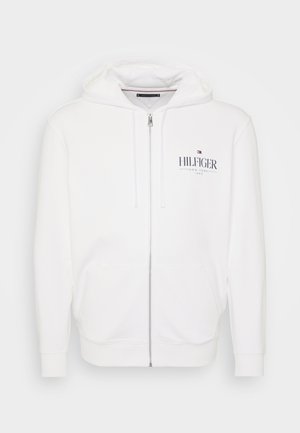 Haut zippé blanc en coton. Présente un logo sur le devant, deux poches, une capuche avec cordon de serrage, et des poignets et un ourlet côtelés. Texture lisse.
