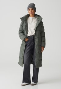 Olijfgroene pufferjas met capuchon, volumedesign, ritssluiting en zijzakken, gedragen over een licht sweater en donkere wijde jeans.