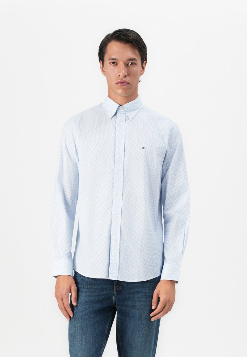 Mann iført en lyseblå skjorte med subtilt mønster og blå jeans, stående mot en ensfarget hvit bakgrunn.
