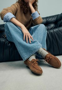 Bruine suède moccasin-stijl schoenen met dunne bruine veters, gedragen met grijze gebreide sokken en lichtblauwe wijde jeans.