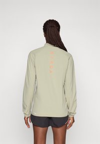 Light green athletic jacket with long sleeves, zip collar, and orange "HYROX" text on the back. Soft, smooth fabric. Worn with black shorts.