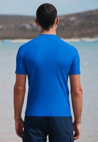 Kortærmet, blå, svedtransporterende skjorte med en glat tekstur og let buet kant, båret af en person med ryggen mod kameraet.