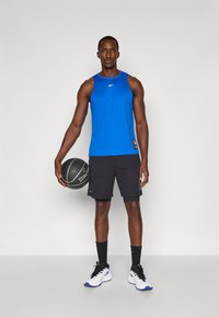Zilā sporta topiņš un melni šorti. Cilvēks tur rokās melnu Spalding basketbola bumbu. Sneakeri ir balti ar ziliem akcentiem.