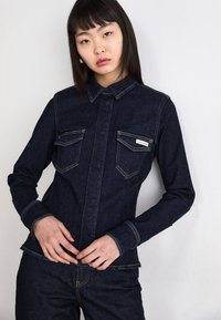 Tmavě modrá džínová bunda s přiléhavým střihem, která má dvě náprsní kapsy, zapínání na knoflíky a kontrastní švy. Hladká textura, dlouhé rukávy.