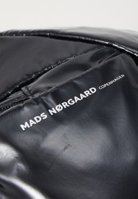 Juodas blizgus pūkinis džemperis su siuvinėta tekstūra, turintis baltą prekės ženklo logotipą, kuris skaito „MADS NØRGAARD COPENHAGEN“ šalia užtrauktuko.