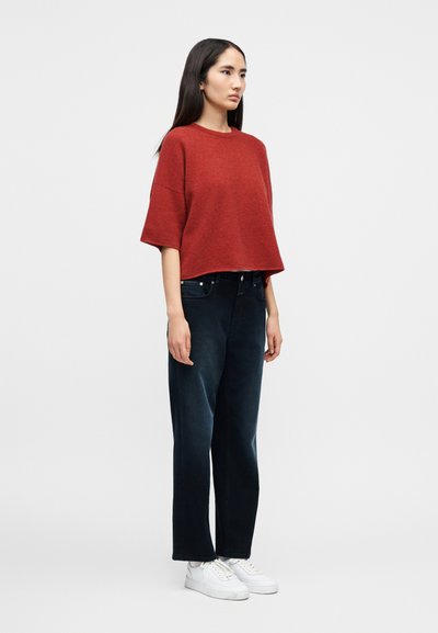 Γυναίκα μ ε μακριά σκουρόχρωμα μαλλιά, που φοράει άνετο κόκκινο πουλόβερ με κοντό μανίκι, σκούρα τζιν φαρδιάς γραμμής και λευκά αθλητικά παπούτσια, στέκεται μπροστά σε ένα απλό φόντο.