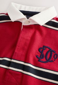 Polo rojo con cuello blanco, con franjas en azul marino y blanco en el pecho y un logotipo bordado en azul marino. Cierre de botones en el cuello.