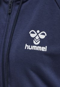 Marinblå zip-up jacka i texturerad tissu. Har en vit logotyp med en abstrakt bi-design, placerad på vänster bröst.