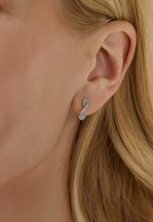 Pendientes de plata con un diseño en bucle, que presentan una superficie texturizada y un pequeño acento brillante. Llevados en el lóbulo de una mujer.