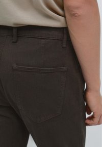 Pantaloni in denim marrone con tasca posteriore di forma rettangolare e cuciture in tono, mostrati da un angolo laterale con parte di un top beige visibile.