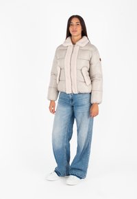 Giacca a vento grigia chiara con colletto peloso e dettagli, chiusura frontale con zip, due tasche laterali, indossata con jeans azzurri e sneakers bianche.
