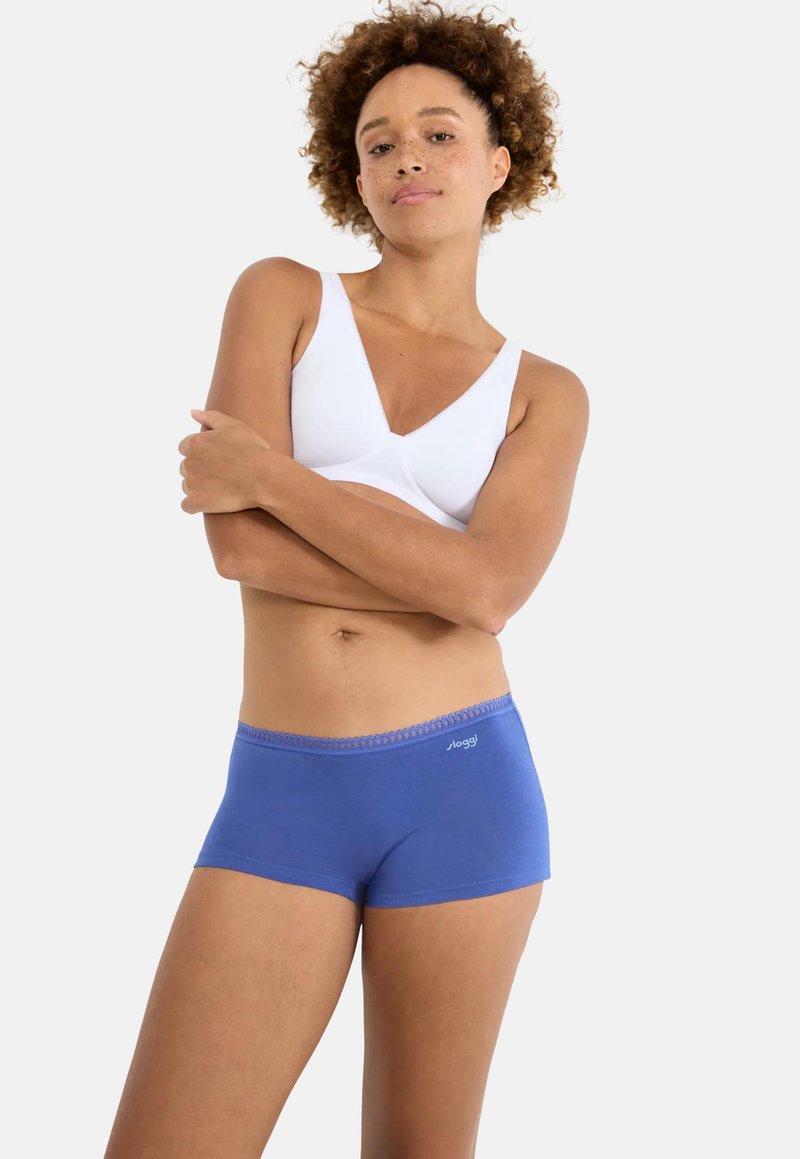 Γυναίκα με σγουρά μαλλιά, φοράει λευκό σουτιέν και μπλε σλιπ τύπου boyshort της Sloggi, στέκεται με σταυρωμένα τα χέρια πάνω σε απλό ανοιχτόχρωμο φόντο.