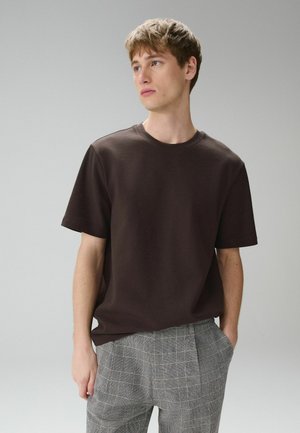 Young man with short light brown hair wearing a dark brown short-sleeve shirt and gray plaid pants, looking sideways with hands in pockets.