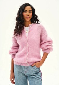 Žena s dlhými kučeravými vlasmi, ktorá má na sebe mäkký ružový sveter na gombíky cez bielu košeľu a svetlomodré džínsy s ružovým bočným detailom.