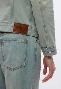 Ljusblå jeansjacka med en brun läderetikett med texten "Calvin Klein" och "CK"-logotypen. Jackan har knäppta ärmslut och kontrastsömmar.