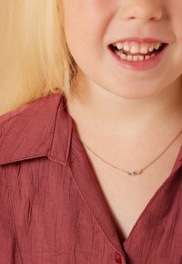 Zilveren ketting met kleine kleurrijke edelstenen, gedragen door een kind in een gemarmerde bordeauxrode blouse met kraag. Close-up van het bovenlichaam en gezicht.