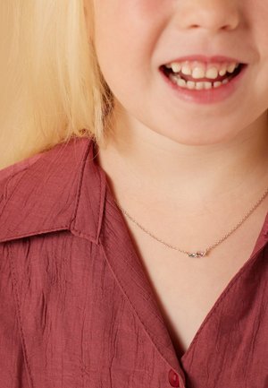 Zilveren ketting met kleine kleurrijke edelstenen, gedragen door een kind in een gemarmerde bordeauxrode blouse met kraag. Close-up van het bovenlichaam en gezicht.