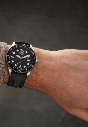 Schwarze Taucheruhr mit silberner Lünette und Markierungen, schwarzem Gummiband, Datumsanzeige, getragen am linken Handgelenk mit beigem Pulloverärmel.