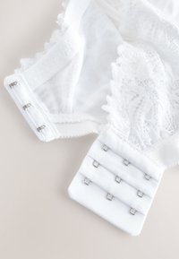 Weißer Spitzen-BH mit transparentem Mesh-Stoff, der drei Reihen von Metallhaken- und Ösenverschlüssen vor hellem Hintergrund zeigt.