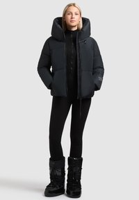 Svart puffjacka med stor huva, dragkedja framtill och sidofickor. Buren över en ribbad svart polotröja och matchad med svarta snöskor.