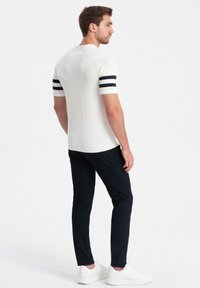 Kortärmad krämfärgad polo med marinblå ränder på ärmarna, kombinerad med mörka byxor och vita sneakers, sedd från baksidan.