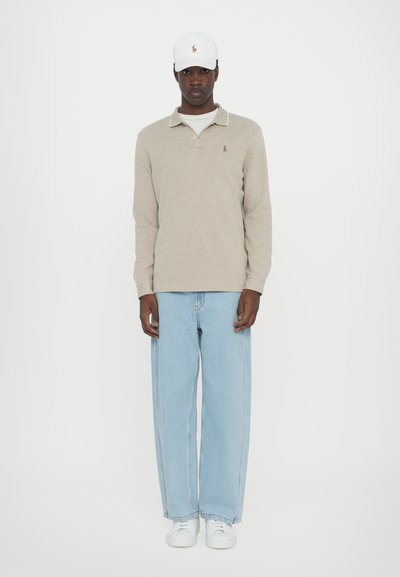 Polo bege de manga longa com colar; jeans de ganga de perna larga azul claro; ténis brancos; boné branco com logótipo à frente; fundo simples.