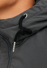 Gray hooded jacket with a zip closure, textured fabric, and black drawstring. Features a silver metal eyelet for securing the drawstring.