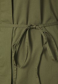 Tessuto verde khaki con una trama liscia, dotato di una cintura legata in vita e pieghe, che creano un aspetto strutturato e aderente.