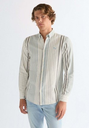 Młody mężczyzna ubrany w jasno-niebieską i szarą koszulę w pionowe paski oraz jasno-niebieskie dżinsy, stojący na tle jednolitego białego tła.