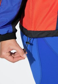 Červená a modrá sportovní bunda s černými detaily; ruka upravuje černý stahovací šňůrku na lemu; texturovaná látka a přiléhavý střih.