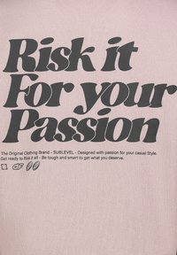 Helles rosa T-Shirt mit großem schwarzem Text, der "Risk it For your Passion" lautet. Enthält Markendetails, die in kleinerem Text darunter gedruckt sind.