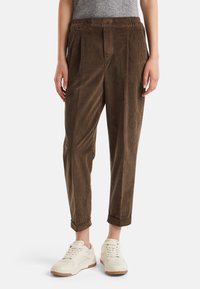 Bruine corduroy cropped broek gedragen met witte sneakers door een persoon die tegen een witte achtergrond staat.