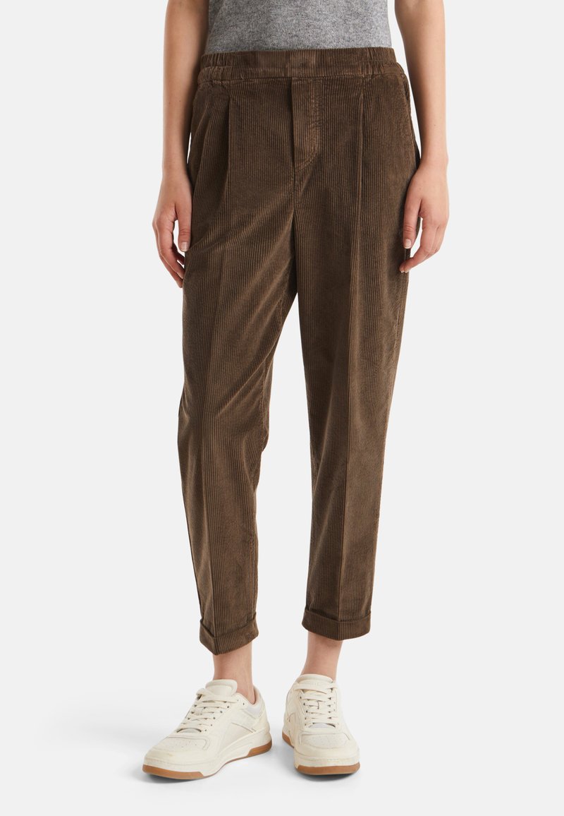 Bruine corduroy cropped broek gedragen met witte sneakers door een persoon die tegen een witte achtergrond staat.