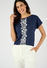 Top de color azul marino de manga corta con bordado floral blanco en el centro, hecho de una tela suave, combinado con pantalones de color claro.