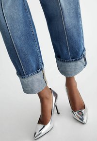 Jeans recortados de color azul claro con puños enrollados, combinados con tacones altos metálicos plateados que tienen una punta afilada y un acabado brillante.