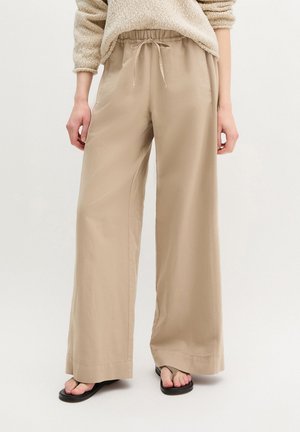 Personne portant un pantalon beige à jambes larges avec une taille à cordon, un pull beige, et des tongs noires, debout devant un fond uni.