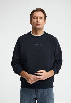 Uomo con capelli castani corti che indossa una felpa nera Polo Club e jeans blu, in piedi davanti a uno sfondo bianco semplice.