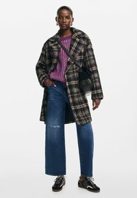 Plaid coat in black, purple, and green, paired with a purple striped sweater and wide-leg blue jeans. Black handbag and sneakers complete the look.