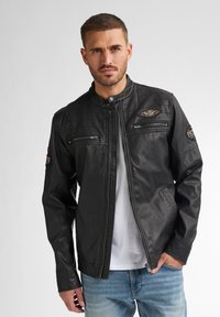 Schwarze Lederjacke mit Stehkragen, mit Reißverschlusstaschen und gestepptem Muster an den Schultern; verfügt über Patches an den Ärmeln.
