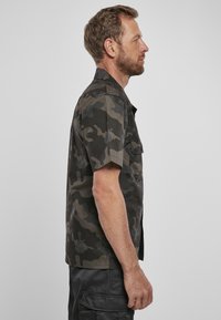Camicia a maniche corte in motivo mimetico scuro, realizzata in tessuto leggero, presenta una chiusura con bottoni e due tasche sul petto.