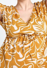 Sukienka w musztardowym kolorze z białym, abstrakcyjnym wzorem kwiatowym, dekolt w kształcie V, krótkie rękawy i ściągacz w talii dla regulowanego dopasowania. Lekką tkanina.