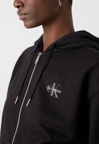 Black zip-up hoodie made of soft fabric, featuring a grey logo embroidered on the chest, with metal zipper and drawstrings.