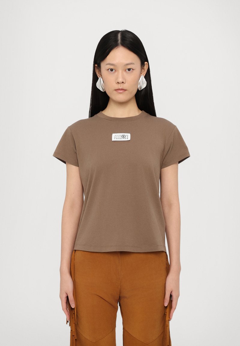 Bruine katoenen T-shirt met korte mouwen en een ronde hals, met een wit etiket op de voorkant. Het model draagt bruine suède broek met franjes.