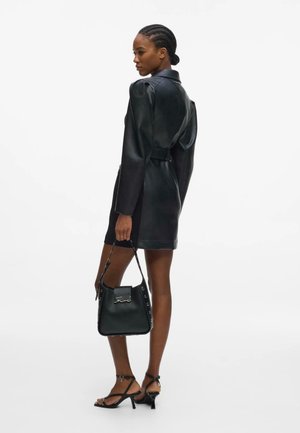 Femme aux cheveux tressés portant une robe en cuir noire, tenant un sac à main noir, et des sandales à talons noires à brides, de profil.
