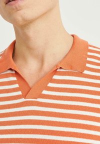 Randig polo tröja i orange och vitt med V-ringning. Tillverkad av ett mjukt stickat material, med krage och korta ärmar.